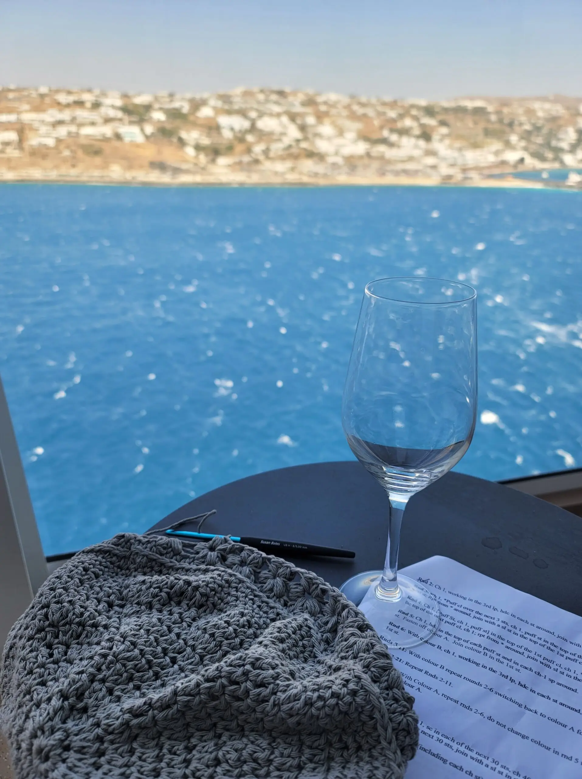 Photo of a coastline in greece in the background, and a table with crochet project, hook, instructions, and empty wine glass in the foreground.
