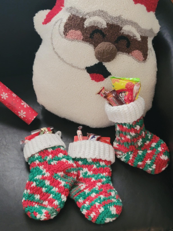 Medium sized holiday stockings in festive red, green, and white yarn with a white cuff with candy inside in front of a santa faced pillow and next to snowflake wrapping paper