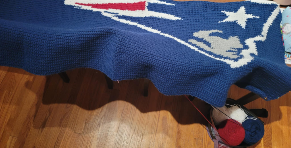 Unfinished logo blanket spread across a table in progress with red, white, gray and blue skeins in a project bag