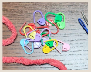 multicolored stitch markers in a small pile next to a strand of yarn and 2 crochet needle heads