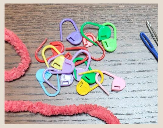 multicolored stitch markers in a small pile next to a strand of yarn and 2 crochet needle heads