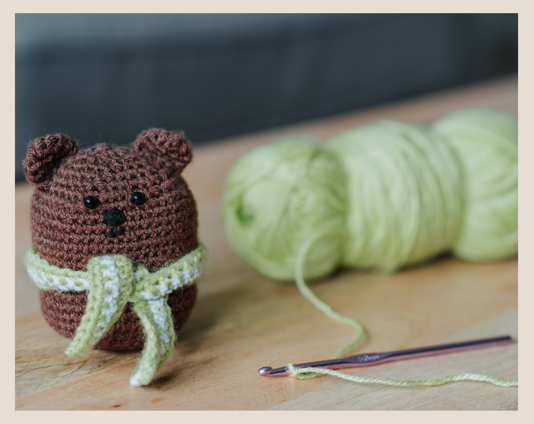 Picture of a brown amigurumi teddy bear with a green skein of yarn to the right of it. There is also a pink crochet hook starting a chain.