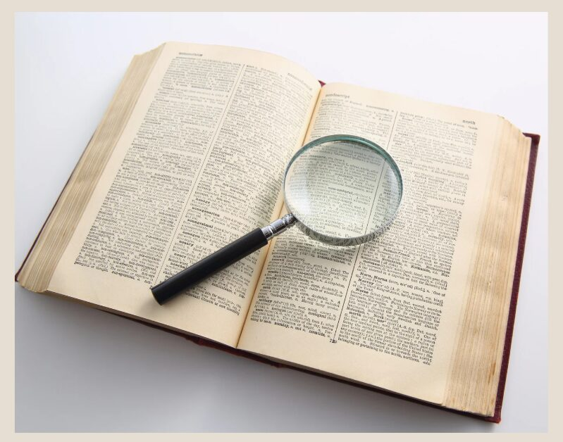 Picture of a white background with an open dictionary that has a magnifying glass laying on top of it.
